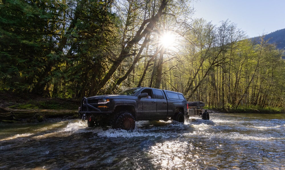 Custom lifted 4x4 truck riding across a river during a sunny day.